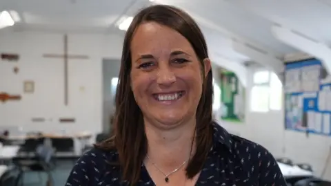 Shaun Whitmore/BBC Anna Price who is looking at the camera and smiling. She is wearing a navy outfit and is standing inside a white painted room.