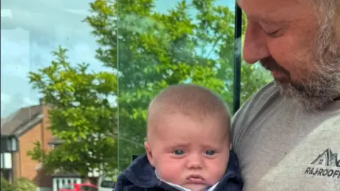 Family A man with a beard and moustache is holding a young baby, which he is looking at with a smile.