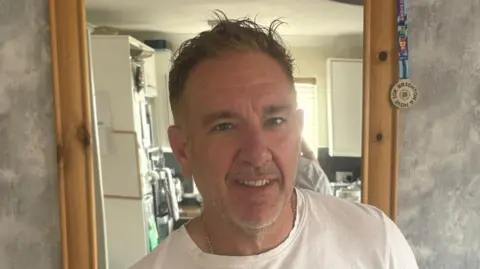 Michelle Fowle Gareth Bowen, who has short hair and is wearing a white T-shirt, smiles in front of a mirror.