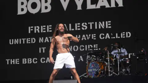 Yui Mok/PA Media A topless man with dreadlocks in white shorts holds a microphone on a stage, with drummer behind. Text on the screen behind reads: Bob Vylan. Free Palestine. United Nations have called it Genocide. The BBC calls it a conflict.