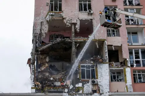 Reuters A pink apartment building with a huge hole in the side of it and a firefighter on a crane spraying water into it.