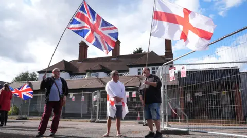 Three men standing outside a large house that is surrounded by metal fencing. The three men are wearing polo shirts, two wearing shorts and two are holding large flags - one is the Union Jack the other is the St George's Cross.