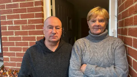 Robby West A man wearing a dark jumper and a woman wearing a grey jumper look towards the camera in front of their home