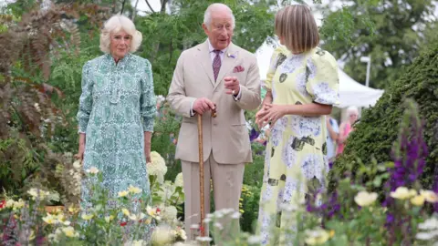 King Charles and Queen Camilla with garden designer Catherine MacDonald are in a garden. Green-leaved trees form the dense backdrop with yellow and purple flowers in the foreground. A pale lemon-coloured hydrangea can be seen too. The Queen is wearing a green-patterned midi dress, MacDonald is wearing a yellow midi dress with a flower print and King Charles is wearing a beige linen suit and is carrying a brown walking stick. 