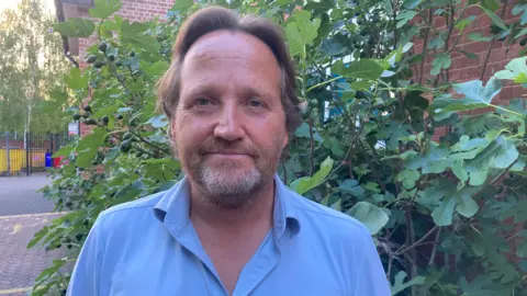 BBC Councillor Matt Dormer is standing outside, wearing a blue shirt. Behind him is a bush, and the road.