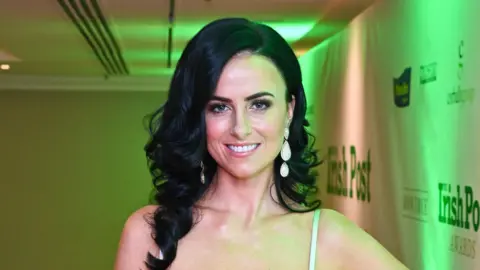 A woman with long black curly hair smiles at the camera as she wears a light coloured strappy dress at an event. She has long pearl drop earrings and is wearing makeup.