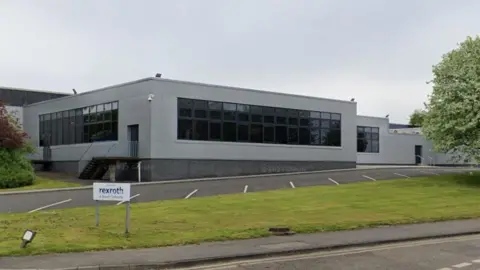 The grey building has lots dark glass windows. It has a flat roof and an empty car park in the foreground with grass and a tree.