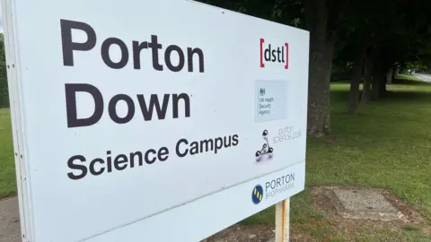 Porton Down Science Campus sign next to some trees with several logos on