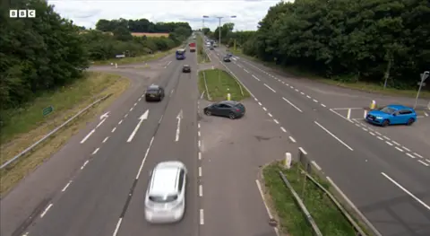 A picture looking down on to the dual carriage way shows a blue car waiting at a junction on the the right side of the road, waiting to cross to a gap in the central reservation where another silver car is preparing to pull on to the the dual carriageway. Multiple cars on the left lane are blurred in the picture as they drive up away from the camera.  