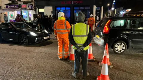 Jozef Hall/BBC Two workmen can be seen and a person in a hi-vis vest are stood near a line of cones. Two cars are being let through the line of cones. People can be seen lining the road in the background. 