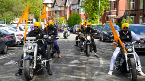 City of Wolverhampton Council Two lines of men in yellow turbans riding motorcycles with orange flags on them bearing the Sikh religious symbol. They are on a road lined with trees and houses one one side. 