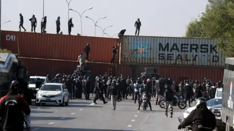 A multi-lane highway with four shipping containers in the middle of it. People are climbing over the containers while police watch on