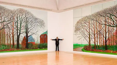 Getty Images Artist David Hockney stands with two very large paintings in an art gallery, depicting colourful trees and houses in the Yorkshire village of Warter.