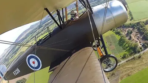 An image recorded by a camera fixed on the wing of the plane shows Tim Rayner in one of the test flights. A carpet of green fields is spread out on the ground.