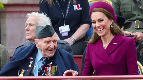 A veteran smiles while wearing military uniform and badges while Catherine, Princess of Wales  laughs sitting next to him wearing a maroon suit and hat, while they watch the procession arrive at Buckingham Palace on Monday.
