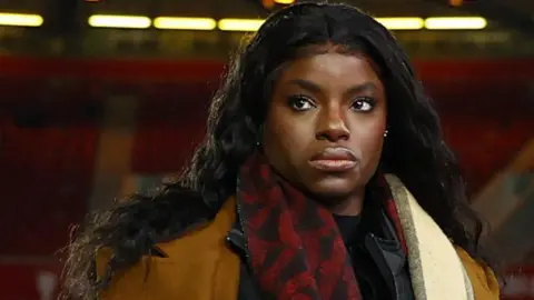 Reuters Eni Aluko, with long black hair and wearing a red and black scarf, brown overcoat and black jacket, looks up and over the camera in a football stadium