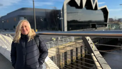 Michaela Ditrichova looking directly at the camera. She has blonde, shoulder-length hair and is wearing a dark blue jacket. She has her hands in her pockets and is standing on a bridge in front of the Glasgow Science Museum. It is a sunny day with a few clouds on a blue sky.