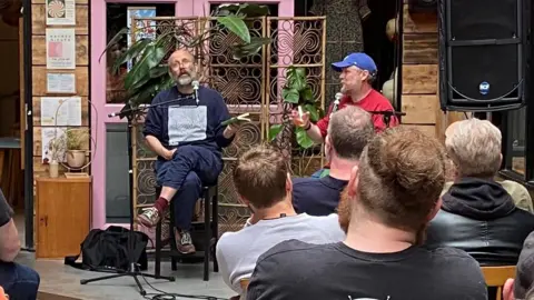 Daniel Kitson, a bald man with a beard, sat besides Tim Key, a man with a beard wearing a blue cap, on stage
