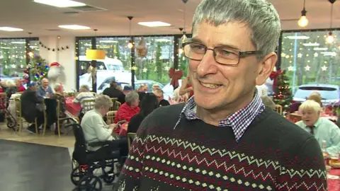 Peter Johnson is standing in a canteen area wearing a blue, white and red checked shirt with a green Christmas jumper on. He is smiling and has glasses on. he has short grey hair.