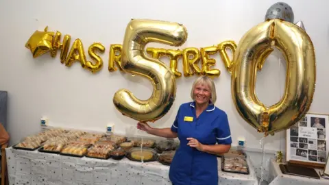 Royal Wolverhampton NHS Trust Julie is wearing her uniform and is stood in front of balloon letters spelling out that she has retired as she holds the numbers 5 and 0 in front of cakes and various treats.
