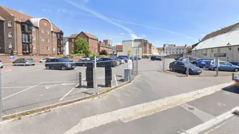 A Google Streetview picture of the car park, which is surrounded by other buildings and has metal fencing around the front of it.