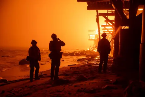 David Crane/Los Angeles Daily News Petugas pemadam kebakaran terlihat siluet di langit kuning saat mereka memadamkan api dari tepi pantai di sepanjang Pacific Coast Highway di Malibu.