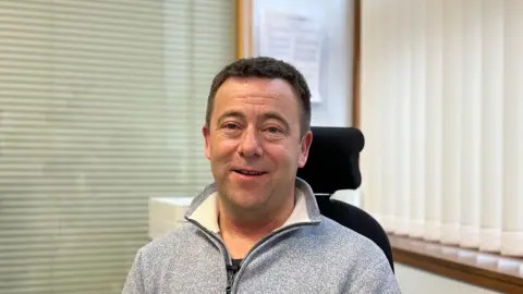 Gary Tumelty sat in his office. He is looking at the camera and smiling. He's wearing a grey quarter-zip jumper.