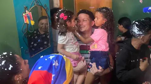 Gustavo Ocando Mervin Yamarte trzyma dwoje dzieci i uśmiechy. Otacza go krewni, z których jeden nosi bandanę w kolorach flagi wenezuelskiej. Mały pokój jest zatłoczony
