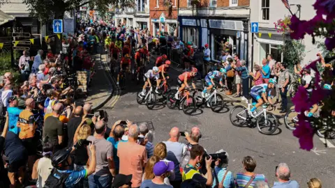 A large group of cyclists riding through a town with crowds cheering them on either side of the street. Some people are taking pictures of the cyclists.