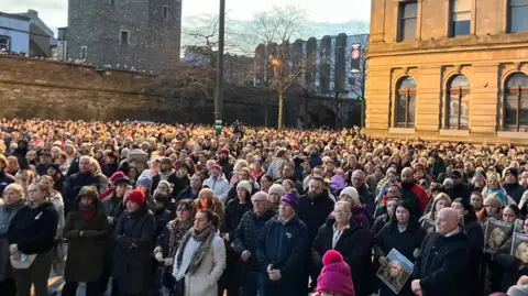 A large crowd of people stand side by side in Derry's Guildhall Square, a number are holding posters of a young woman.