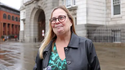 Jack Valpy/BBC A woman in a coat outside a large stone building. She is looking off to the side of the frame.