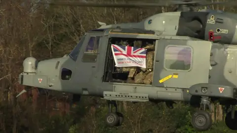 A helicopter flying above a field with military personnel waving a Union Jack.