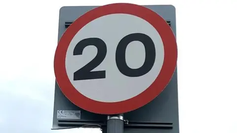 A road sign depicting a 20mph speed limit. It is circular in shape, the 20 is written in black writing on a white background, with red trim.