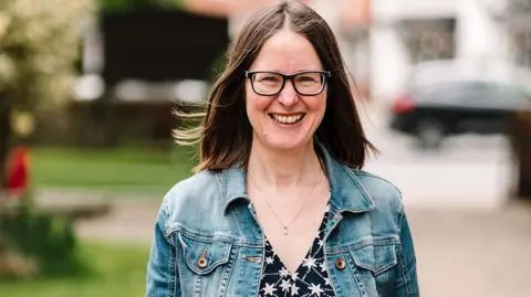 Visit Knowle A woman with long brown hair, glasses, a denim jacket and black top with white stars, smiles as she walks towards us from the road.