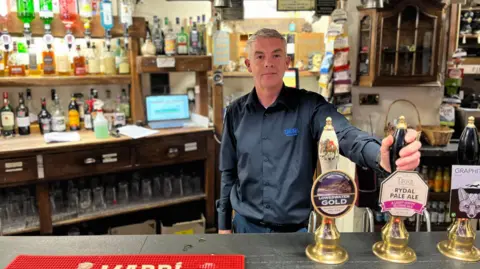 John Cook standing behind the bar in the Sun. He has short grey hair and is wearing a black shirt.