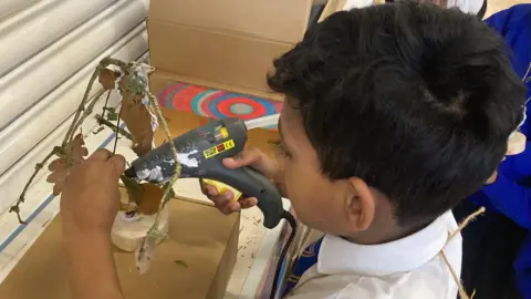 Ryan is Nine and is holding a hot glue gun to attach parts to his sculpture. He is wearing a white shirt with a blue and yellow Camp Hill Primary School tie.