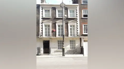 Front view of a 18th Century brick townhouse in London. It has a black front door and appears to be three storeys tall. There is a traditional black lamppost on the pavement outside