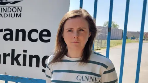 Councillor Kate Tomlinson stands in front of blue railings looking stern. She has mid-length brown hair and is wearing a white and blue striped top that says "Cannes" on it.