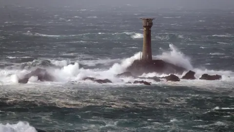 PA Media Longships Lighthouse is being blasted with waves whipped up by winds on a sunny day. 