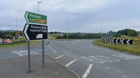 A roundabout with a green sign saying Welshpool and a black sign saying Maesbury Road industrial estate. White road markings can be seen on the roundabout.