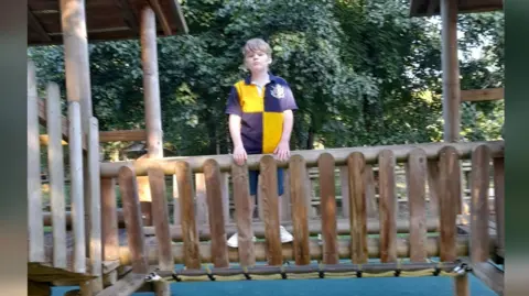 Victoria Dean Alex posing for a photograph on the wooden bridge on a climbing frame in a playpark. He is wearing a yellow and blue quartered rugby style top and is looking at the camera. He is not smiling.