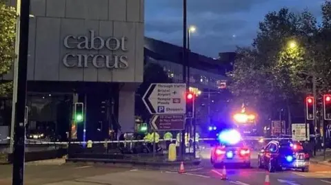 Cabot Circus shopping centre at night. There are several police cars parked next to it with their flashing lights on, and a police cordon is in place at the scene of the crash.