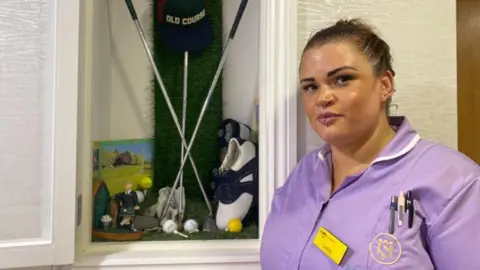 BBC/Angela Ferguson A woman with tied back brown hair and wearing a lilac coloured uniform with a name badge saying 'Hello my name is Candice' is smiling as she stands next to a display of golf clubs, golf balls, an ornament of a man playing golf and some golf shoes.