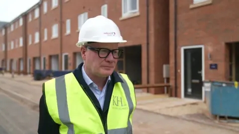 Richard Parker wearing a white builder's hat, glasses and a hi-vis jacket over a suit, stands and looks to our right. Behind him is a row of homes that are under construction.
