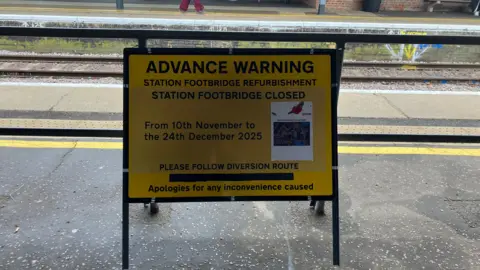 Vikki Irwin/BBC A yellow sign sits on a platform. It gives an advanced warning of footbridge refurbishment works and the dates they will take place. 