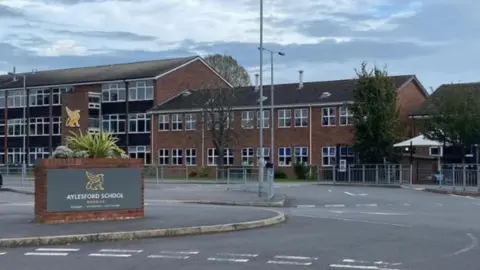 Handout Buildings, including one featuring three storeys, are in the background behind railings and a road. In the foreground, a brick wall near a road includes the words Aylesford School.