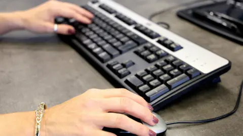 BBC Woman's hands with dark pink nails, using a computer keyboard and mouse.