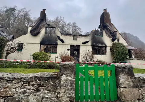 Jason O'Flynn A burnt out cottage. The roof is gone, there is red and white tape in front of the building behind a green gate