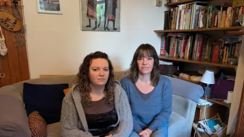 Alex Moss/BBC News A picture of Juliet Rimer with long, curly, brown hair and wearing a black top and grey cardigan. She is sat on a sofa next to a bookshelf alongside Kate Rimer, who has long, brown hair and is wearing jeans and a blue jumper.