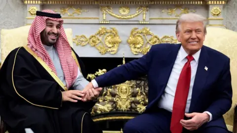 Reuters Mohammed bin Salman and Donald Trump sit side-by-side in front of a gold-embossed fireplace in the Oval Office. The two men are both smiling and Trump is holding out his right hand which the prince is clasping.